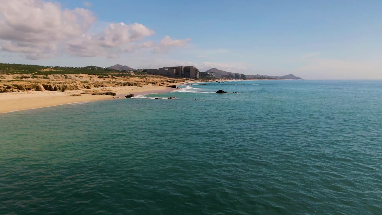 At 0:02 in El Tule Los Cabos, the turquoise water spreads across the lower frame while the beach curves past dark rocks toward distant buildings and hazy hills, with no visible people.