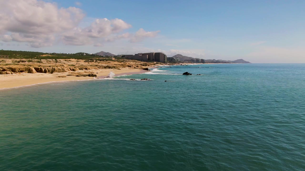 At 0:03 in El Tule Los Cabos, a brighter line of white surf breaks onto the sandy beach while calm blue-green water, shoreline rocks, dunes, distant buildings, and hills remain visible with no people in frame.