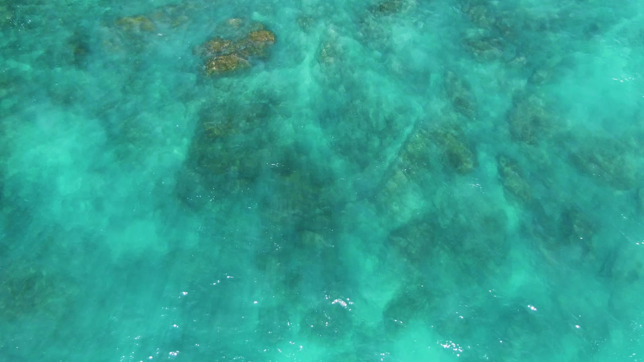 Straight-down view of clear turquoise water at El Tule Los Cabos with scattered dark seabed formations near the upper portion and no visible people.