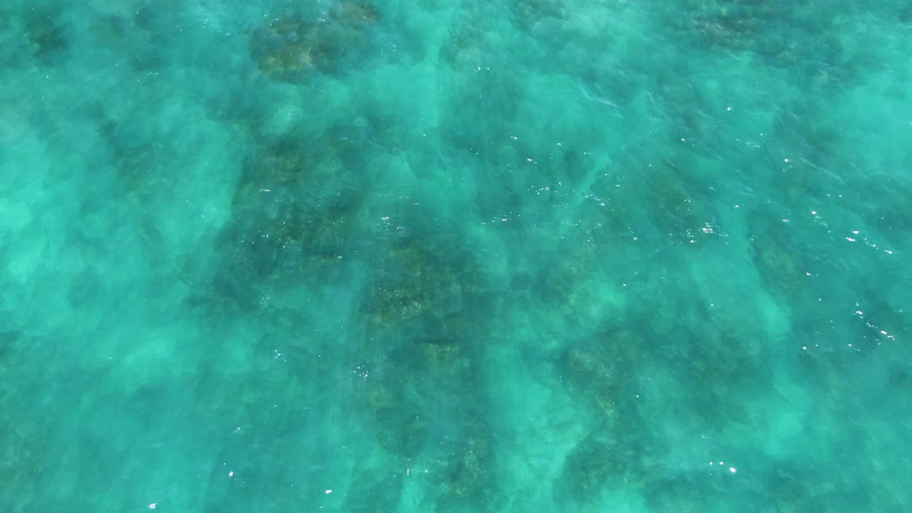 Turquoise water at El Tule Los Cabos fills the frame as ripples pass over a mottled seabed with darker patches concentrated toward the center and upper edge, with no visible people.
