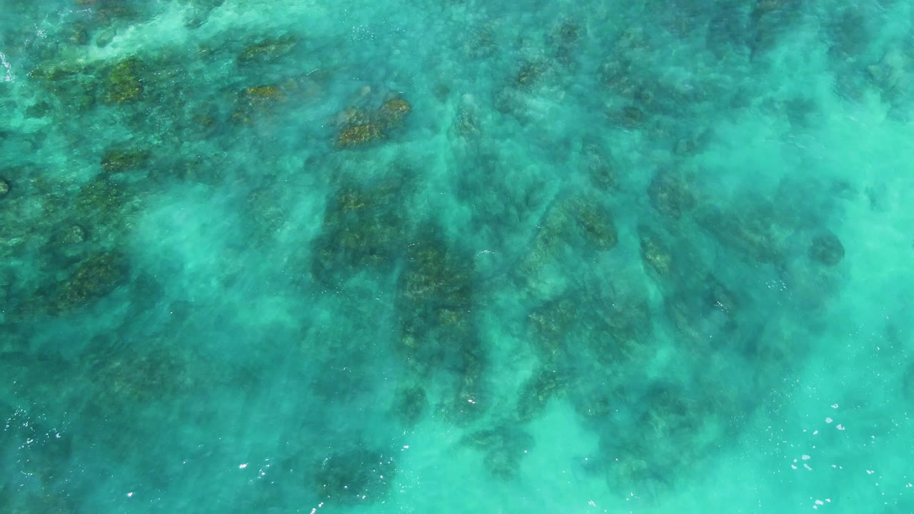Aerial-looking frame of shallow turquoise ocean at El Tule Los Cabos, where irregular rock shapes sit below a lightly rippled surface with no visible people.