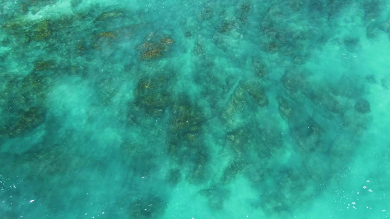 Turquoise water at El Tule Los Cabos shows underwater rocks spread across the frame, with small bright ripple marks near the lower edge and no visible people.