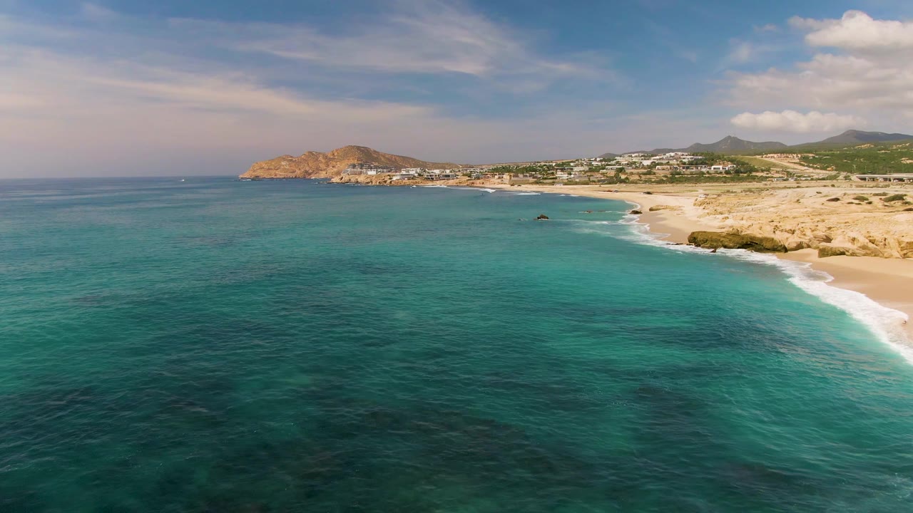 Aerial view at El Tule Los Cabos with turquoise water in the foreground, a pale sandy shoreline to the right, rocky coastal points, distant hills, and no visible people.