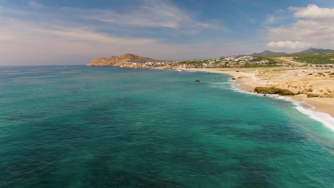 At 0:01, El Tule Los Cabos shoreline curves across the frame with deeper teal water at left, white foam along the beach, tan rocks near shore, inland buildings, hills, and no visible people.