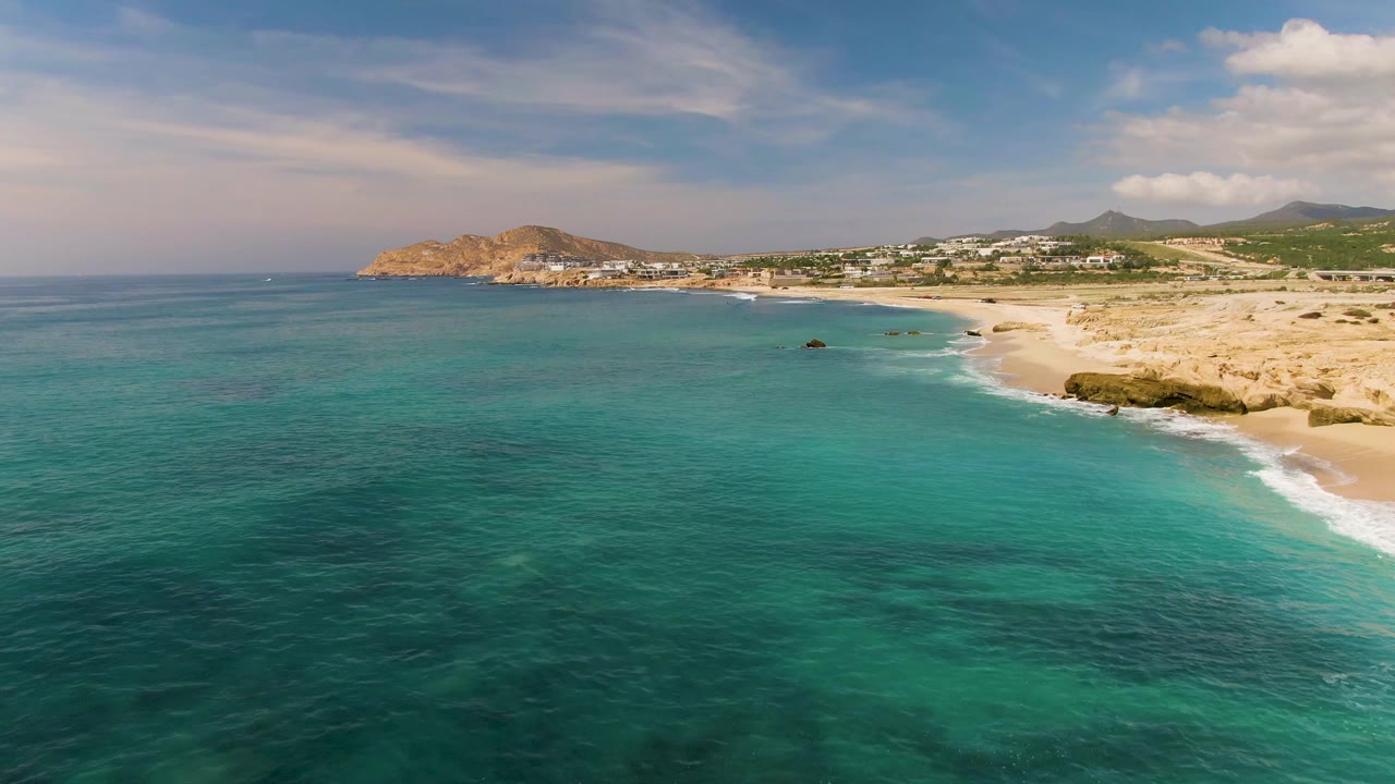 At 0:02, the frame holds more open turquoise water before the El Tule Los Cabos beach, with small dark seabed patches, light surf on the right edge, rocky coast, distant hills, and no visible people.