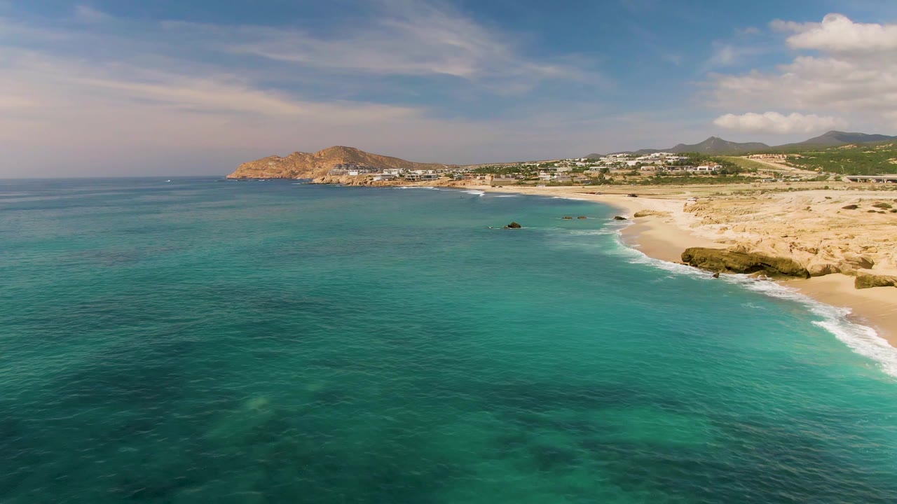 Wide coastal view with teal water, sandy beach, rocks, and distant hills.