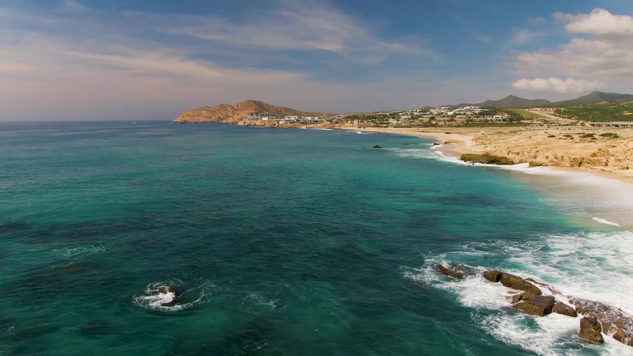 Aerial frame from El Tule Los Cabos showing turquoise ocean in the foreground, white surf around dark rocks, sandy beach to the right, distant hills, and no visible people.