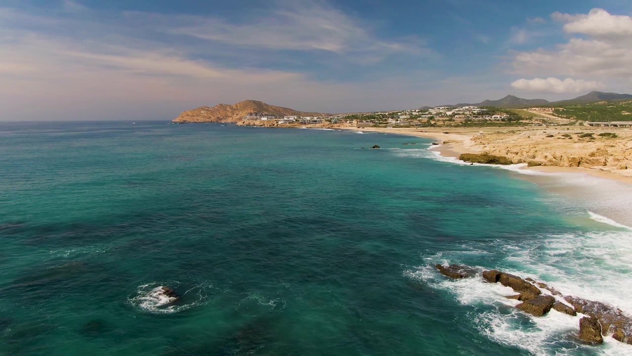 Wide aerial shoreline with waves breaking over rocks beside a sandy beach.