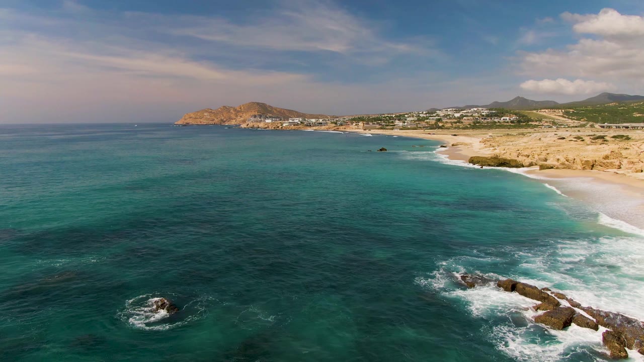 Aerial El Tule Los Cabos frame showing more open sea, a curved surf line along the sand, rocks with foamy water near the lower right, and no visible people.