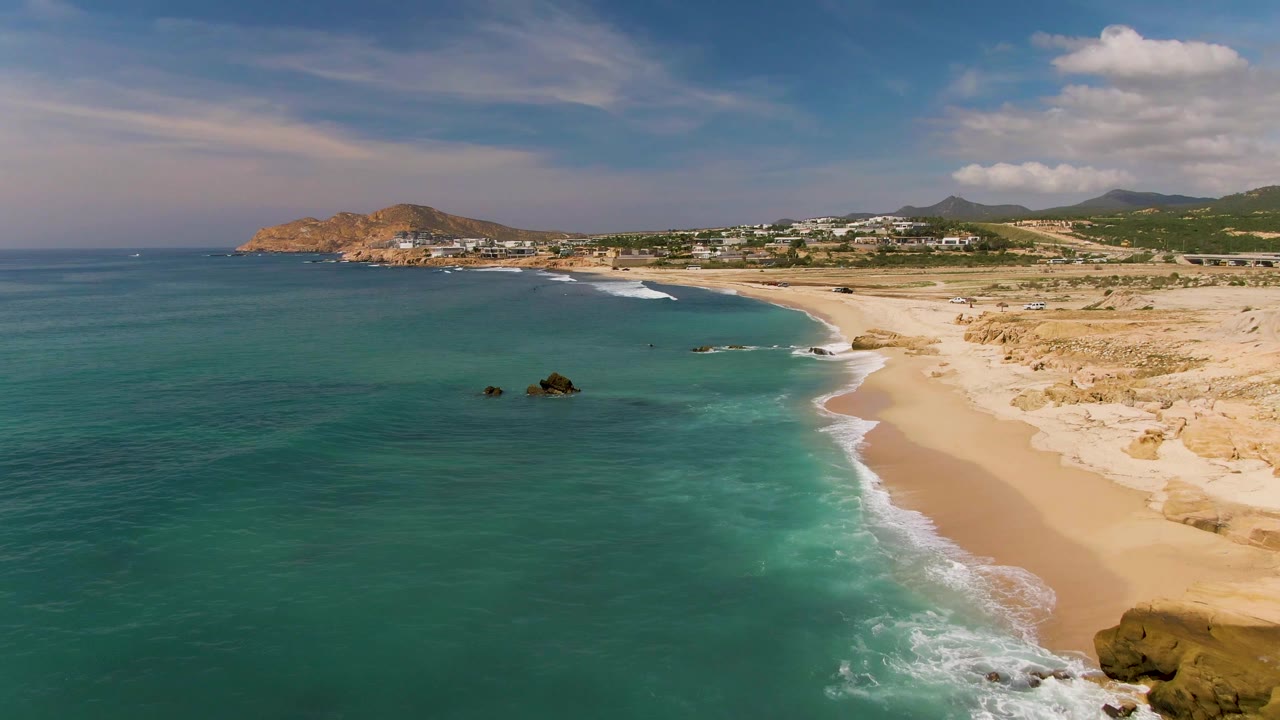 Aerial frame from El Tule Los Cabos showing turquoise water, a sandy shoreline, scattered offshore rocks, distant hillside buildings, and no visible people.