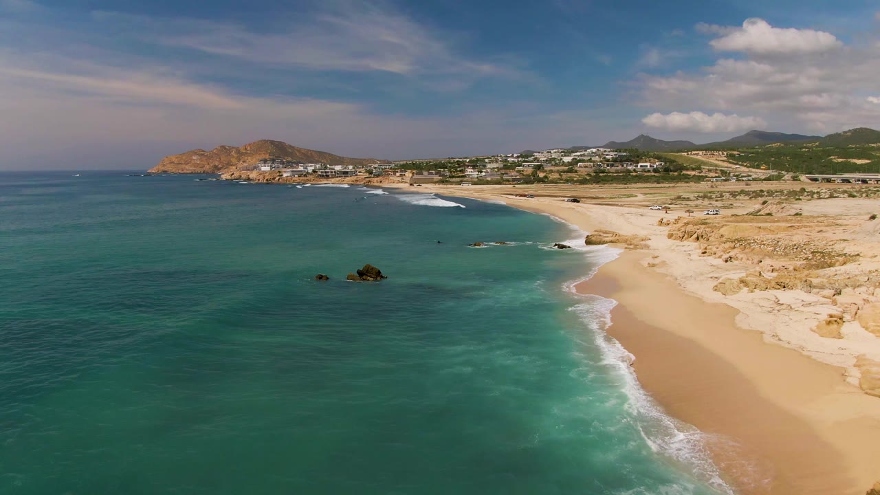 Aerial frame from El Tule Los Cabos with the shoreline angled through the scene, small waves reaching the sand, dark rocks offshore, and no visible people.