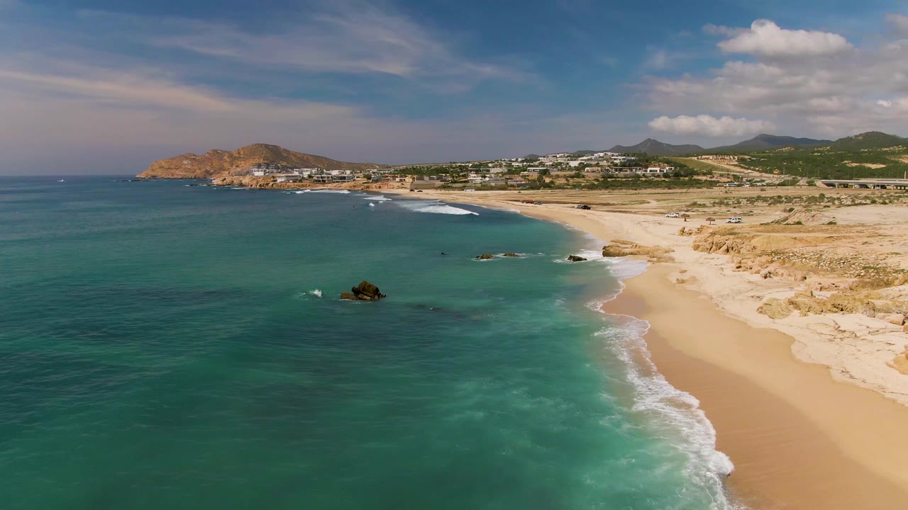 Aerial frame from El Tule Los Cabos showing a wider stretch of empty sand, clear turquoise water, small surf lines, rocky outcrops, distant buildings, and no visible people.