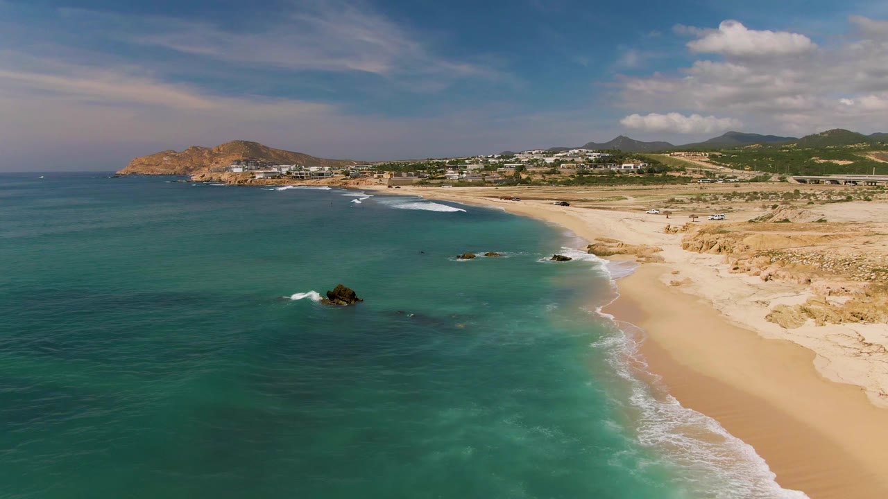 Aerial frame from El Tule Los Cabos where white surf traces the shoreline, dark rocks sit in the water, the sandy beach runs toward distant hills, and no visible people appear.
