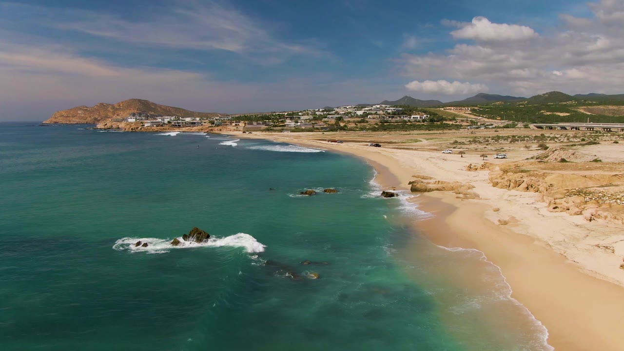 Aerial frame over El Tule Los Cabos with turquoise water on the left, a sandy beach on the right, rocky surf near the foreground, distant hillside buildings, and no visible people.