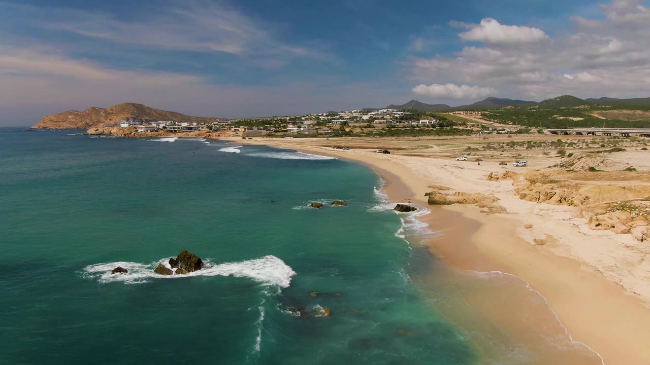 At 0:01, waves spread white foam around the central rocks off El Tule Los Cabos while the empty sand curves toward hillside buildings under scattered clouds.