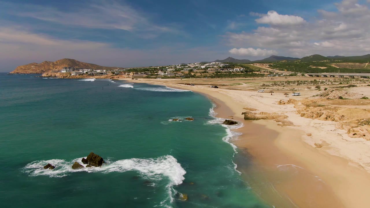 At 0:02, white surf wraps more fully around the offshore rocks, with clear green-blue water in front, broad sand to the right, and no visible people on the beach.