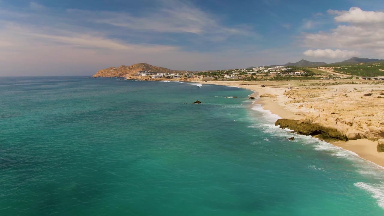 High aerial view over El Tule Los Cabos, Mexico, with turquoise water in the foreground, a sandy beach curving along the right, rocky shoreline at lower right, distant hills, and no visible people.