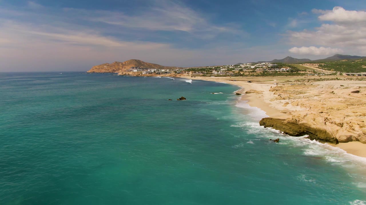Aerial frame over El Tule Los Cabos, Mexico, where turquoise water occupies the left foreground, white surf reaches the sand, tan rocks line the lower right edge, and no visible people are present.