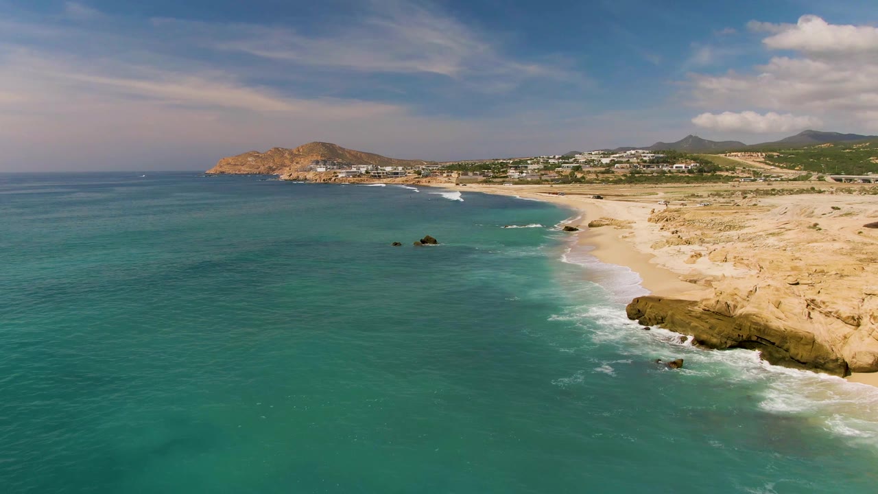 Aerial view of El Tule Los Cabos, Mexico, with a broad turquoise sea, pale beach on the right, small dark rocks offshore, tan rocky ledges in the lower right, distant hills, and no visible people.