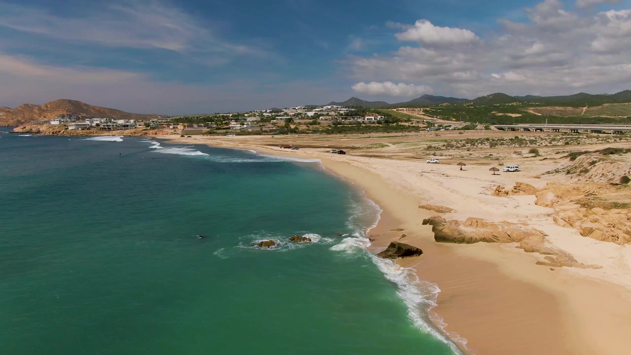Aerial frame over El Tule Los Cabos showing turquoise water on the left, a curving sandy beach, surf around scattered rocks, distant hills, and small vehicles onshore.