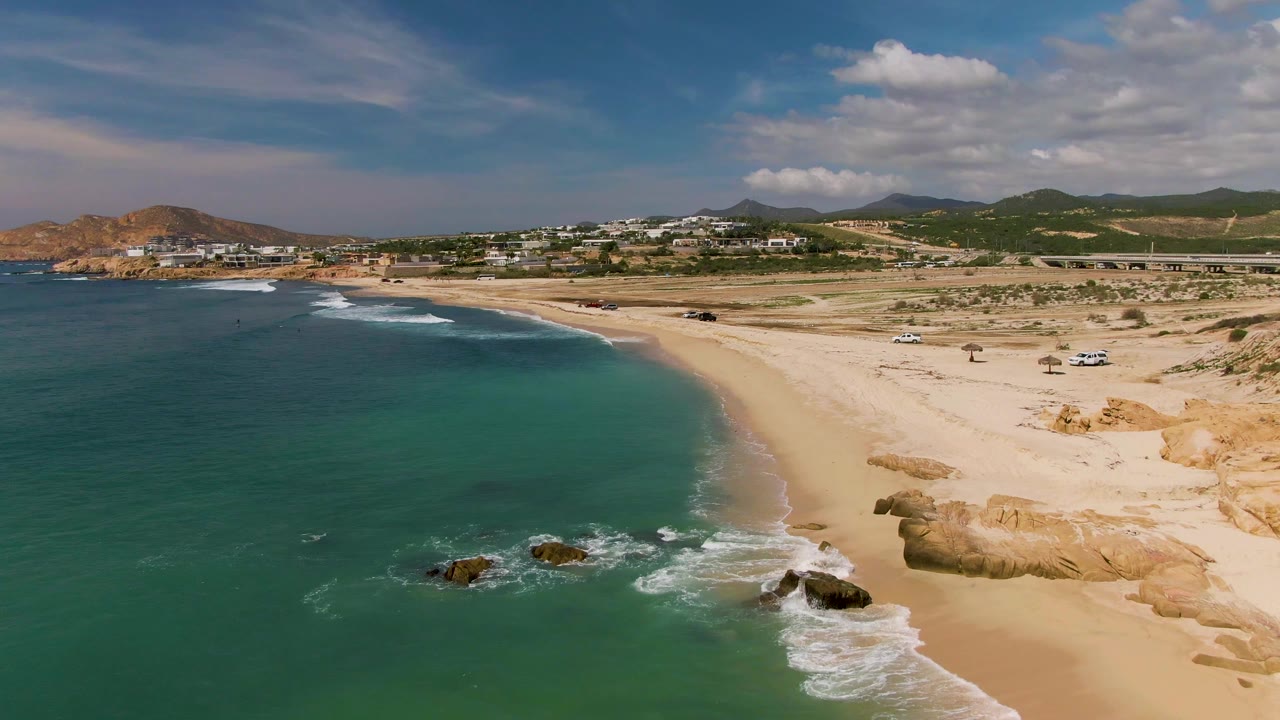 El Tule Los Cabos aerial frame with green-blue water across the left side, rocks ringed by white foam near the beach, pale sand and dunes to the right, and tiny beach activity in the distance.