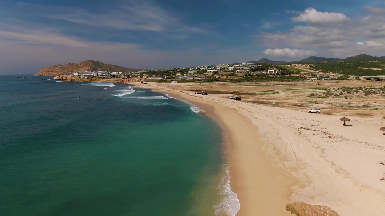 Opening aerial frame over El Tule Los Cabos with turquoise water beside a sandy beach, white surf lines, a few parked vehicles, distant buildings and hills, and no visible people.
