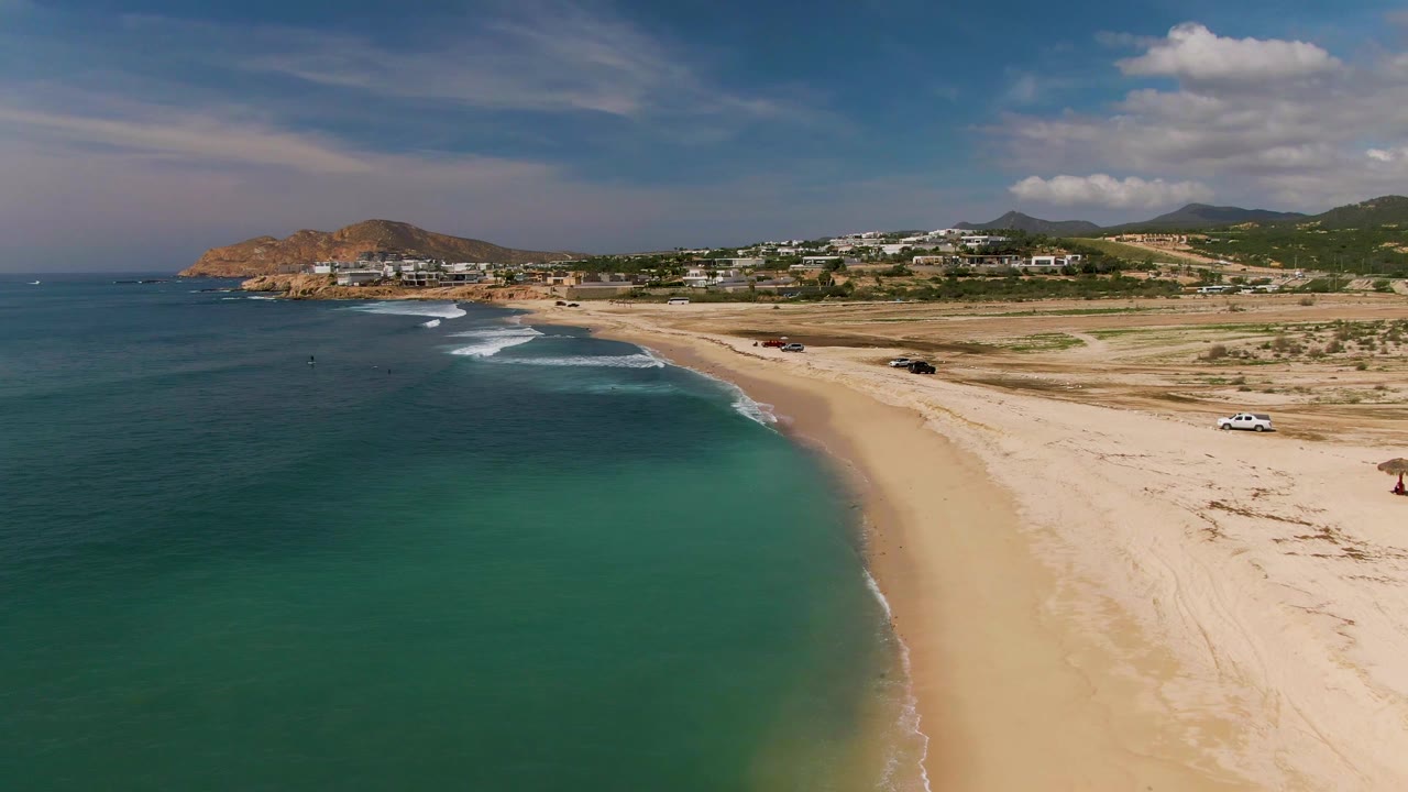 At 0:01, the El Tule shoreline curves through the frame with green-blue water, foamy waves reaching the sand, vehicles set back on the beach, distant buildings, hills, and no visible people.