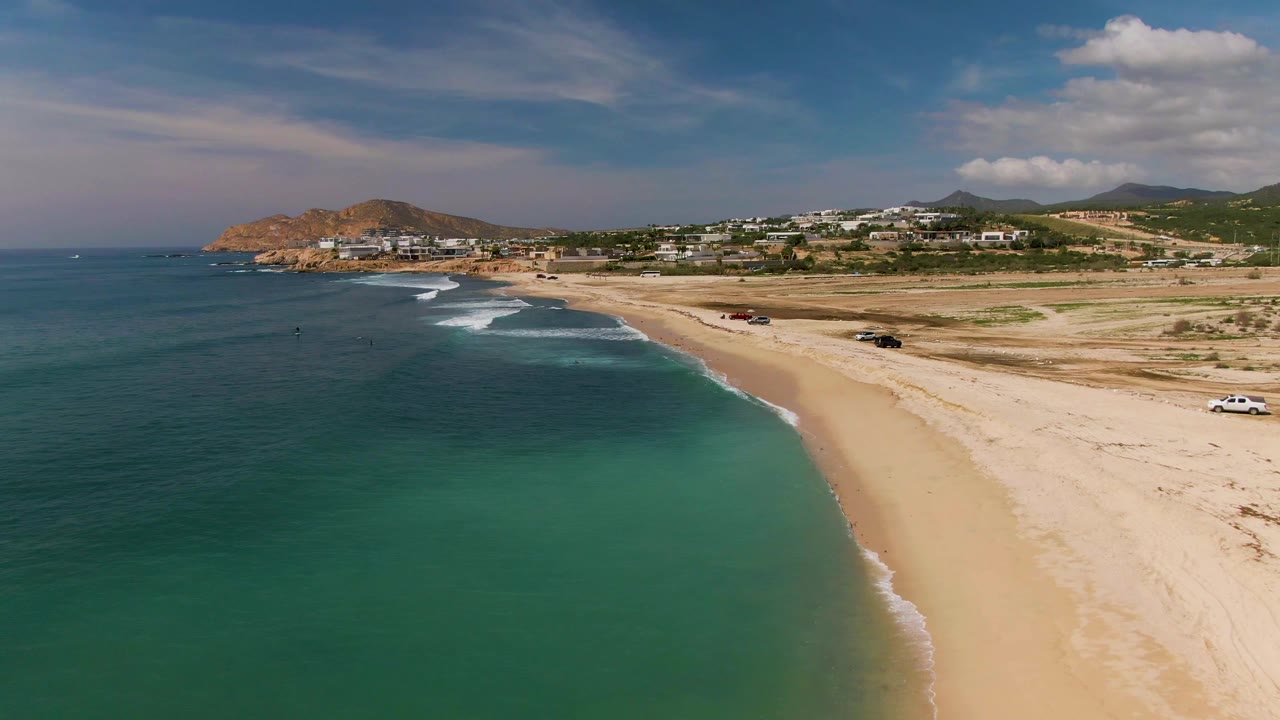 At 0:02, the drone perspective holds wide over El Tule Los Cabos, showing a larger band of turquoise shallows, pale sand, small surf breaks, parked vehicles, background hills, and no visible people.