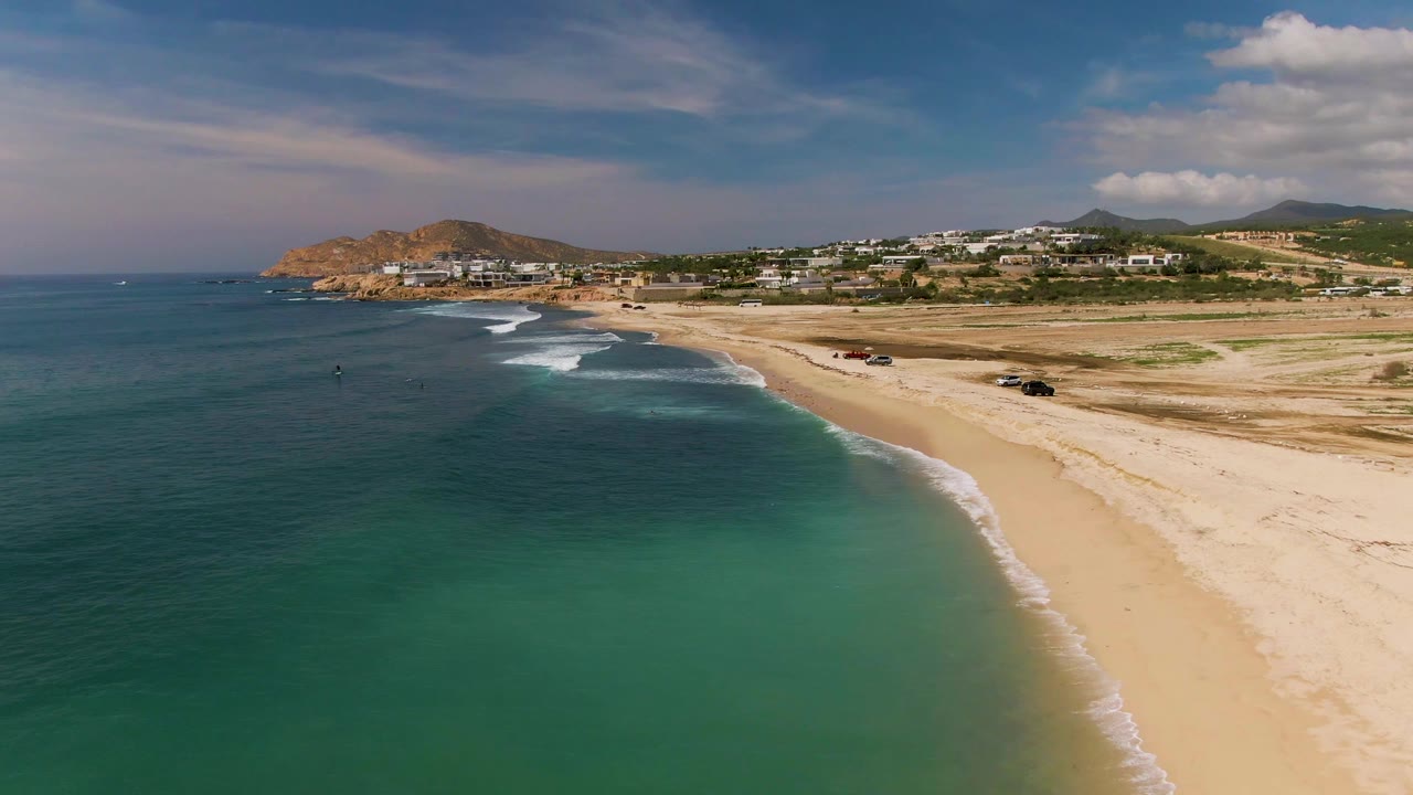 Aerial shoreline with dark blue ocean, green shallows, breaking waves, sand, vehicles, and hills.