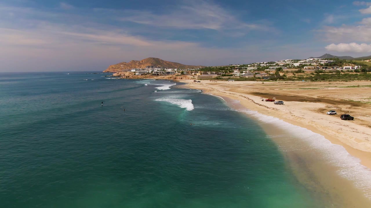 Aerial view over El Tule Los Cabos with teal water in the foreground, a sandy beach at right, parked cars on the shore, low buildings, hills, and small distant people in the water.