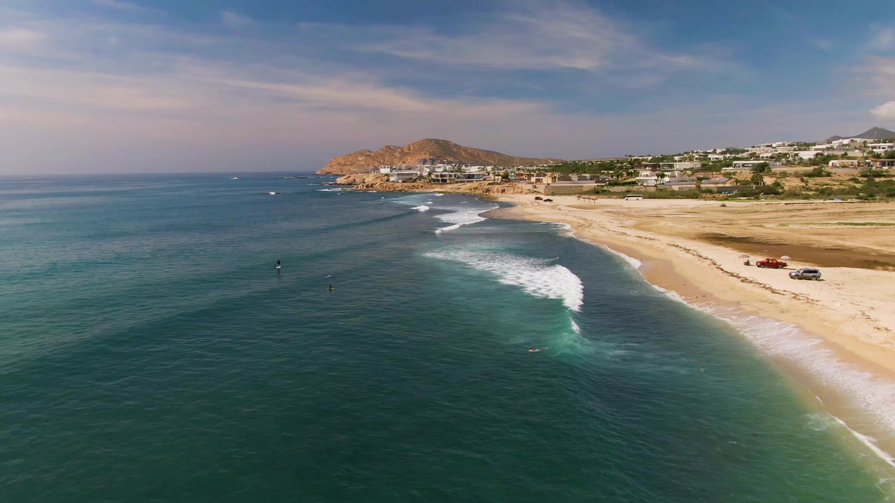 At 0:02, the surf sits closer to the right side of the frame along El Tule Los Cabos, with deep teal water across the foreground, tan sand, beach vehicles, and low buildings under streaked clouds.