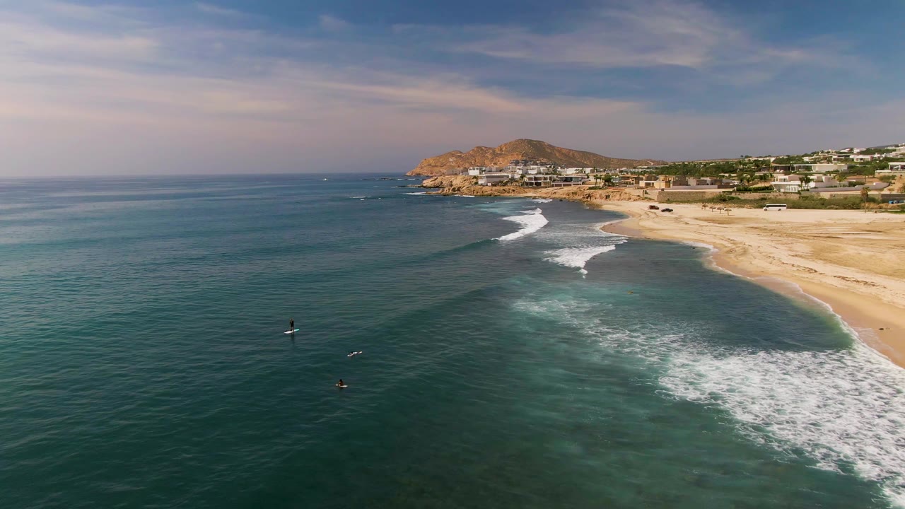 Aerial view over El Tule, Los Cabos with two small board riders in blue-green water, white surf breaking toward a sandy beach, and hillside buildings in the distance.