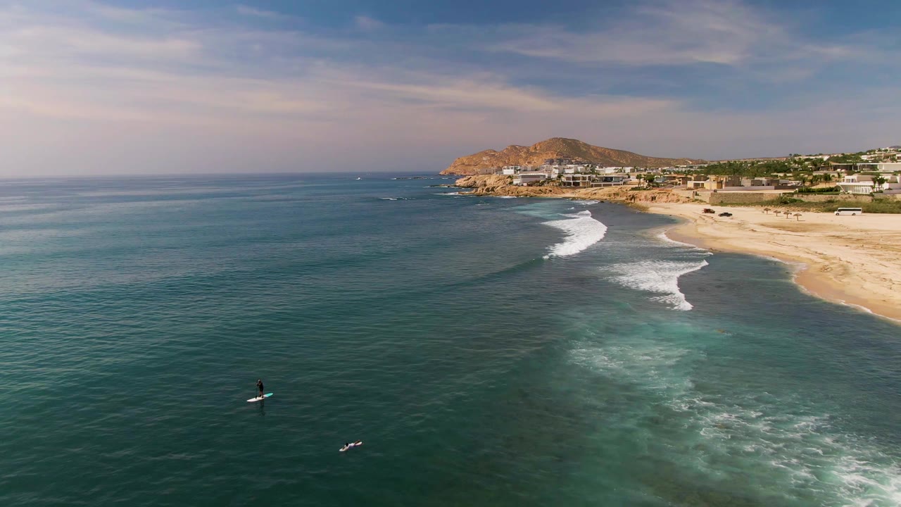 El Tule coastline fills the frame with dark blue water, two small visible board riders, a pale sand beach, white foam bands, and hillside development under a partly cloudy sky.