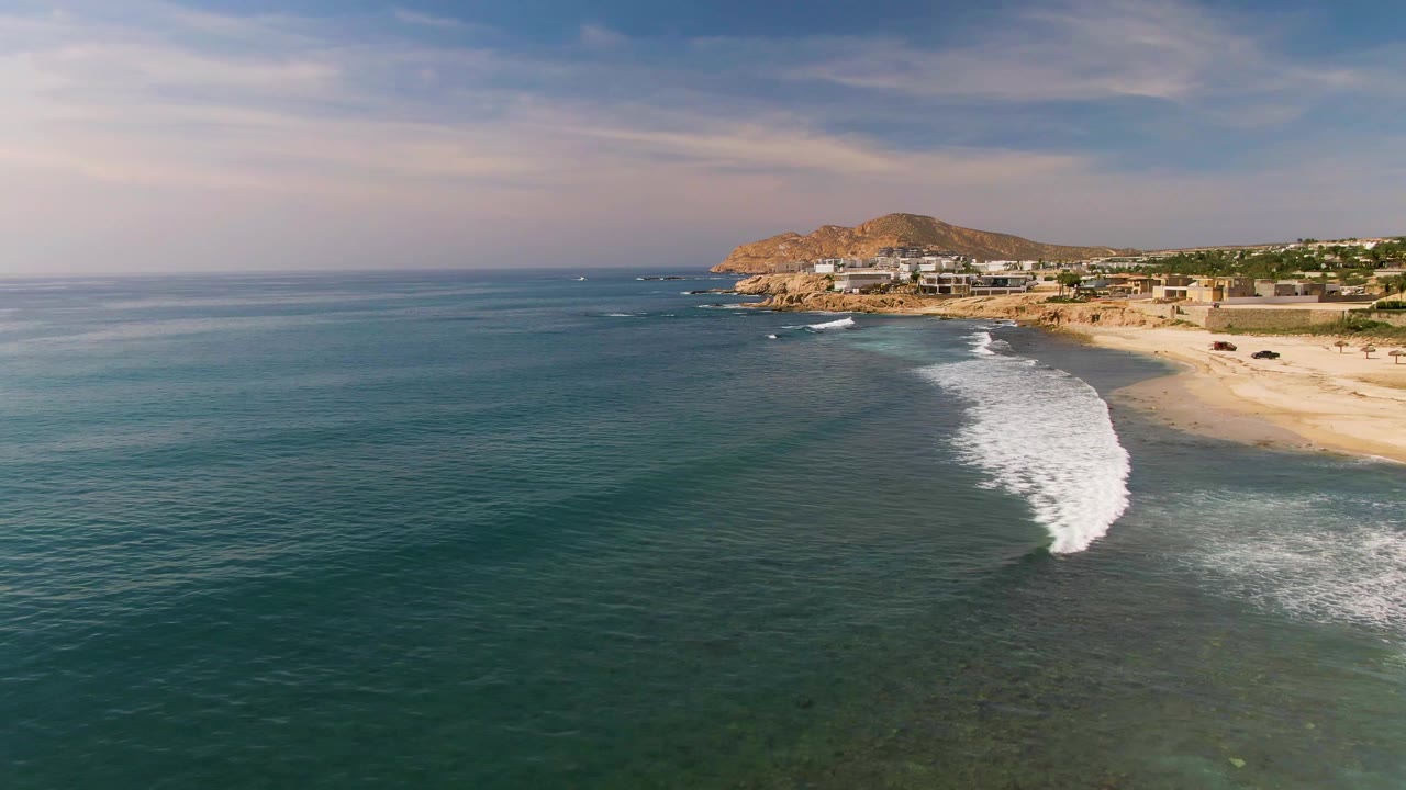 At 0:00, an aerial view over El Tule Los Cabos shows blue ocean, turquoise shallows, a white wave line reaching the sandy beach, coastal buildings, a rocky hill, and no visible people.