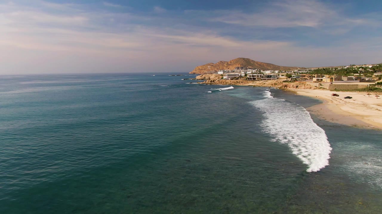 At 0:01, the breaking surf has moved farther through the center of the aerial El Tule Los Cabos frame, with open ocean at left, sandy shore at right, buildings, hill, and no visible people.