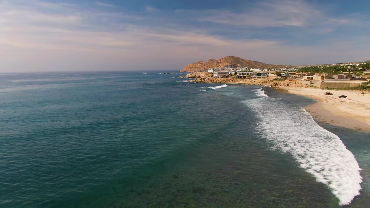 At 0:02, the wave break stretches diagonally along the right side of the El Tule Los Cabos shoreline, with darker water in the foreground, beach and buildings beyond, and no visible people.