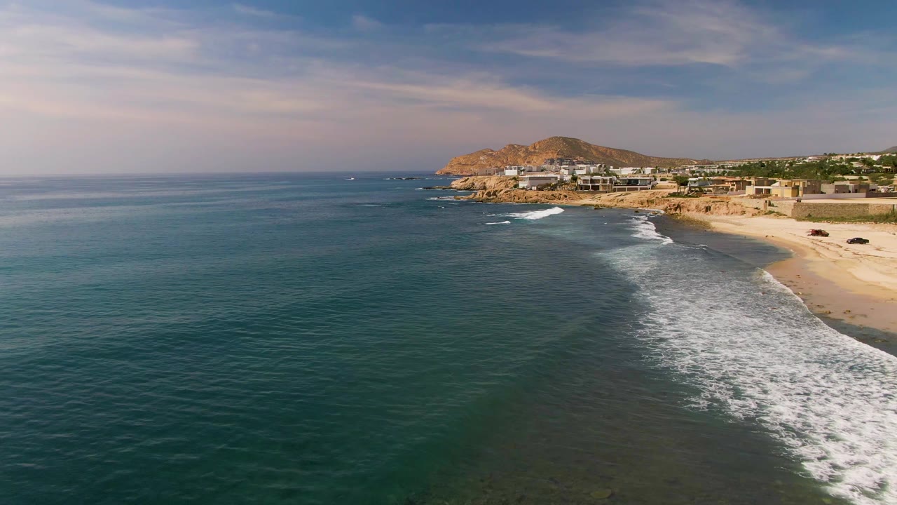 Foamy wave spreads along a sandy beach below coastal buildings and a hill.
