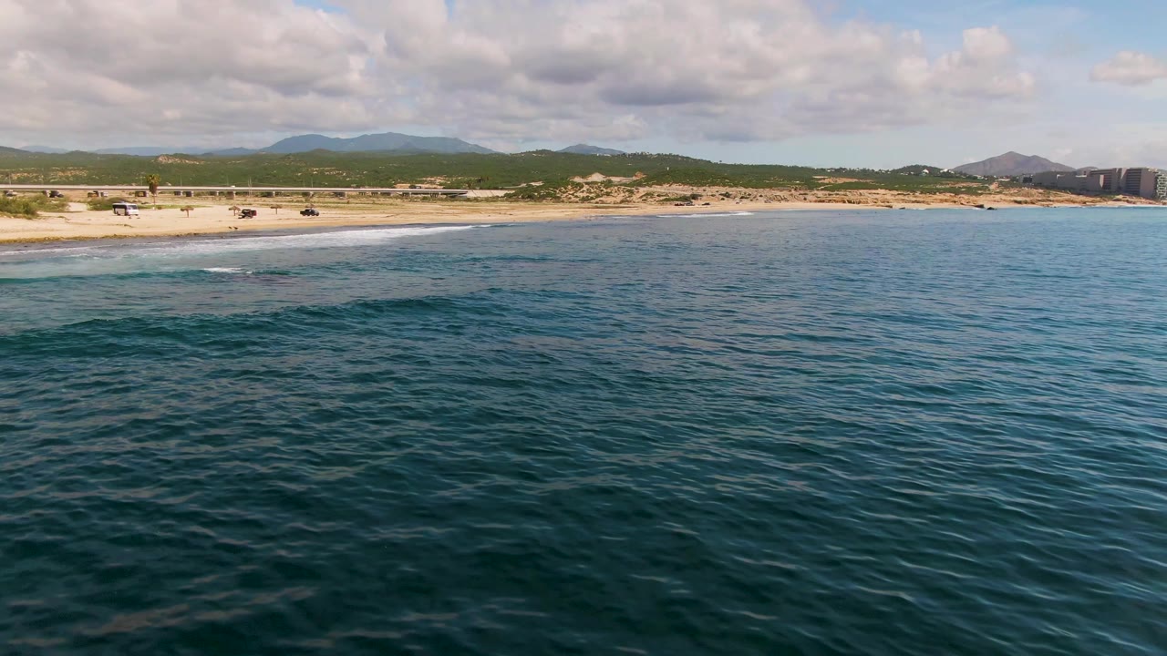 Aerial view at El Tule Los Cabos with blue-green ocean in the foreground, a sandy beach and road beyond it, distant mountains under partly cloudy sky, and no visible people.