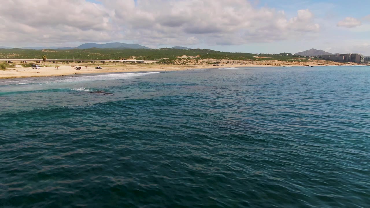 Open ocean with small whitecaps approaching a sandy shoreline and mountain backdrop.