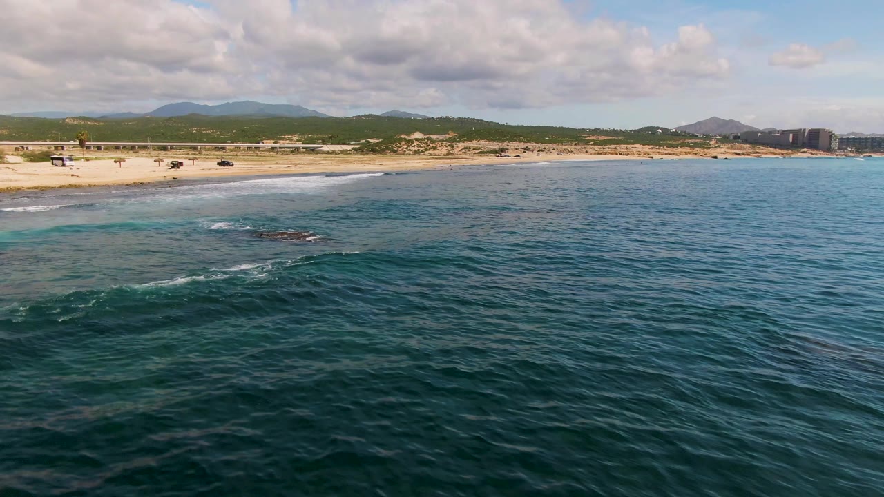 Aerial frame from El Tule Los Cabos showing darker reef or rock patches beneath the water, sandy shoreline, beach road, distant buildings, mountains, and no visible people.