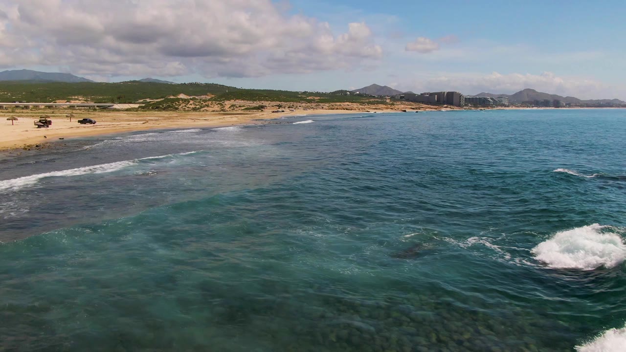 Aerial frame over El Tule Los Cabos showing turquoise water in the foreground, a sandy beach on the left, white surf near shore, distant hills and buildings, and no visible people.