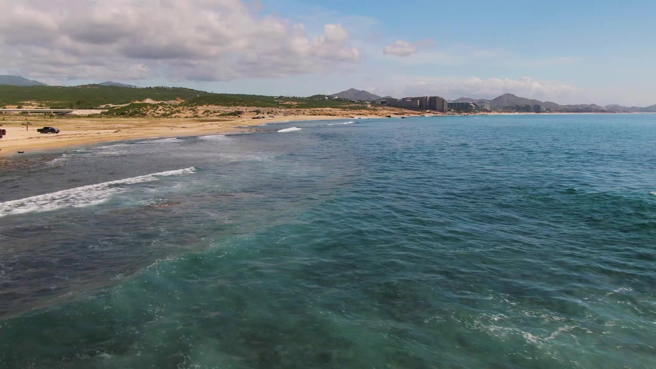 Aerial view at El Tule Los Cabos with the sandy beach along the left edge, low white waves crossing darker shallow water, blue ocean to the right, distant hills, and no visible people.