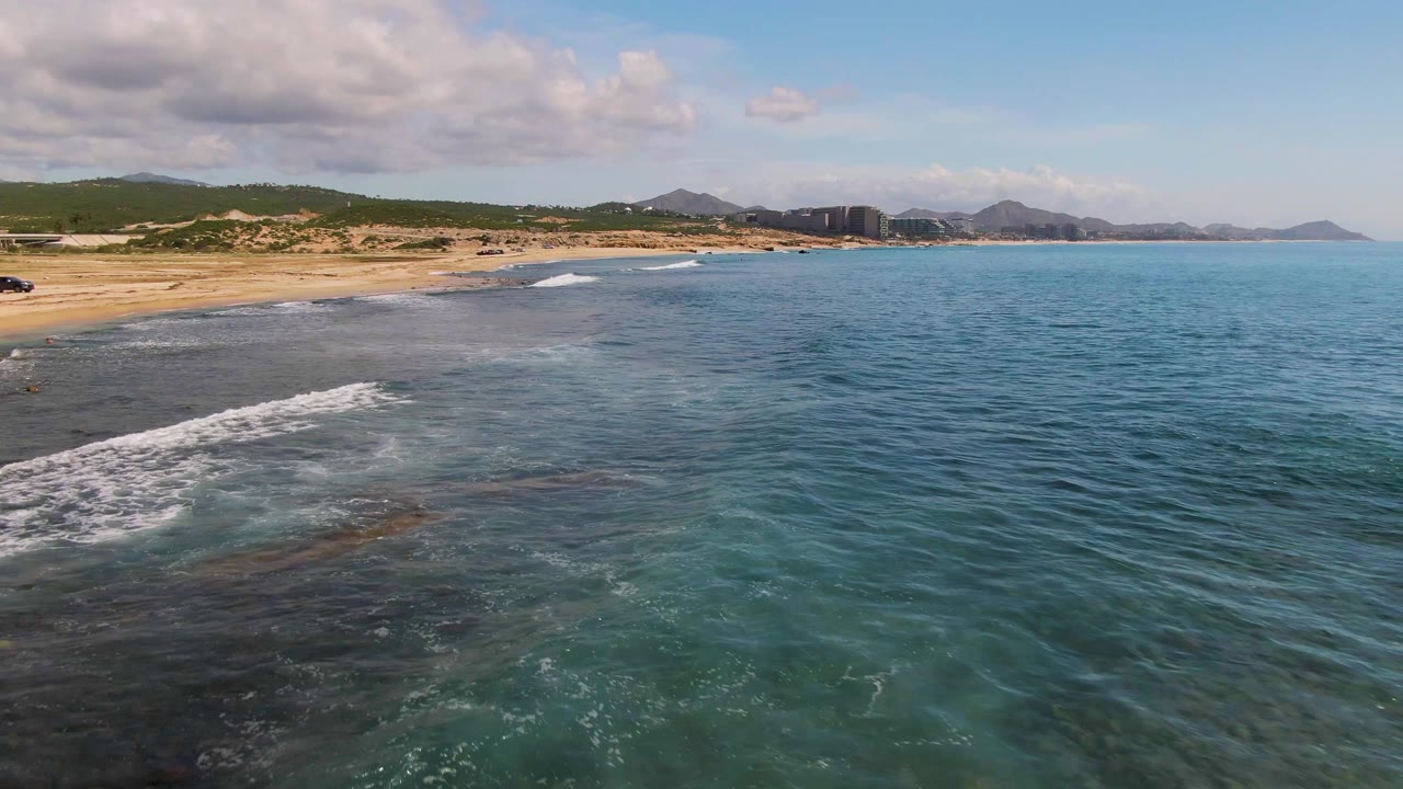 Aerial coastal frame from El Tule Los Cabos showing clear blue-green water, a band of white foam near the beach, sandy shore on the left, distant buildings under hills, and no visible people.