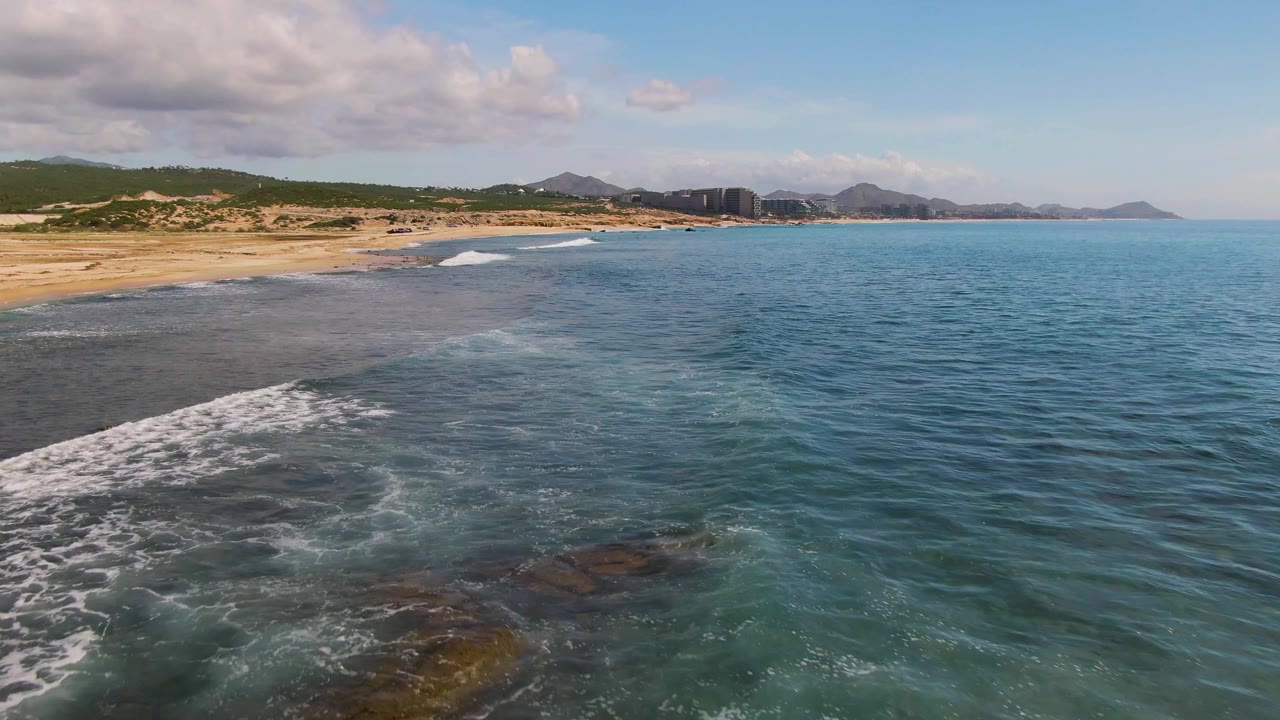 Aerial frame over El Tule Los Cabos with white surf entering from the lower left, turquoise ocean across the right side, sandy beach and dunes along the left, distant hills and buildings, and no visible people.