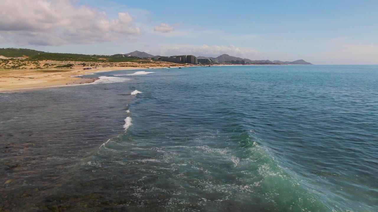 Opening aerial frame over El Tule Los Cabos, Mexico, with blue ocean on the right, sandy beach on the left, small breaking waves near shore, distant hills, and no visible people.