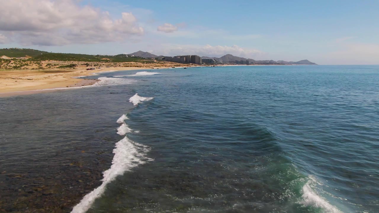 Aerial view at El Tule Los Cabos with a longer white surf line crossing dark nearshore water, pale sand to the left, open blue sea to the right, distant hills, and no visible people.