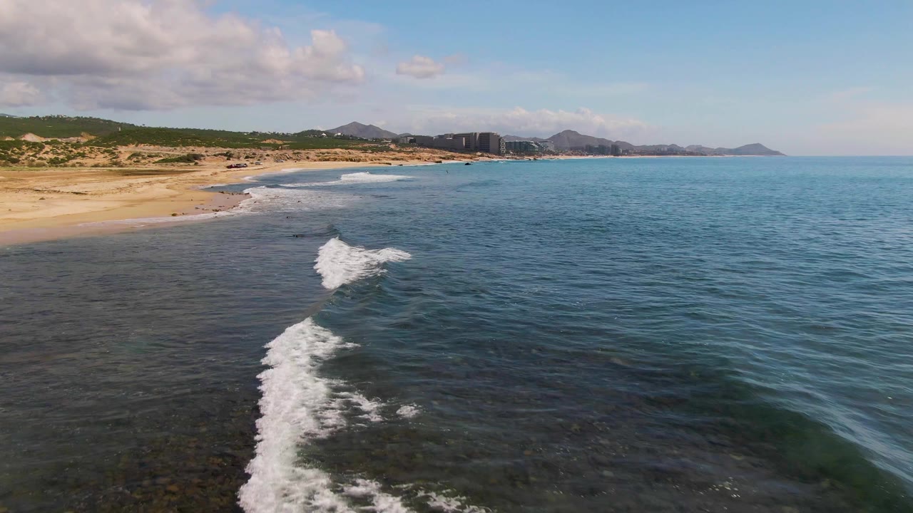 El Tule Los Cabos shoreline seen from above as white foam thickens near the center, with the sandy beach running along the left edge, deep blue water to the right, hills beyond, and no visible people.