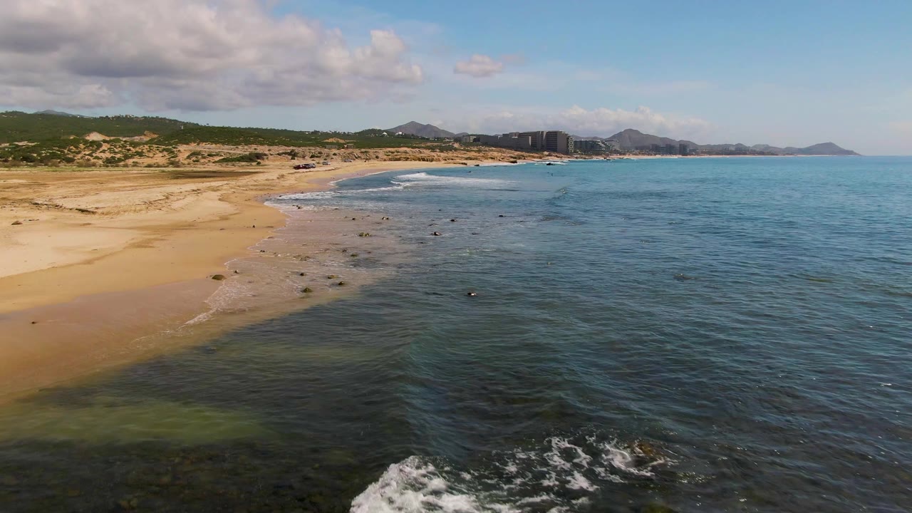 Aerial view of sandy beach, shallow blue water, rocks, hills, and clouds.