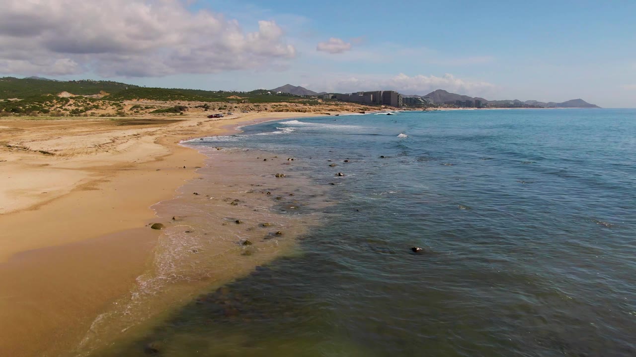 At 0:01, the El Tule Los Cabos shoreline curves across the frame, with wet sand exposed, scattered rocks in shallow water, distant buildings near the hills, and no visible people.