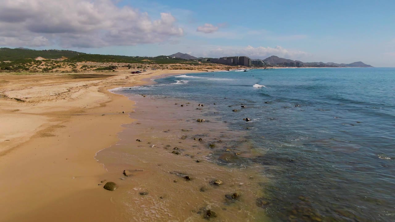 At 0:02, small waves move toward the El Tule Los Cabos beach, with more shallow rocks visible near the waterline, tan sand to the left, and no visible people.